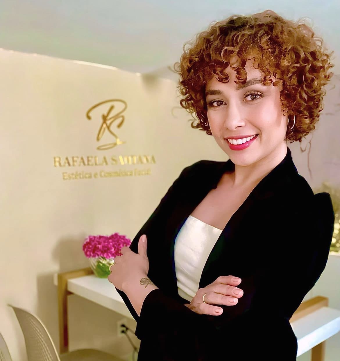 Rafaela Santana de blazer preto na recepção, diante do logo dourado da clínica